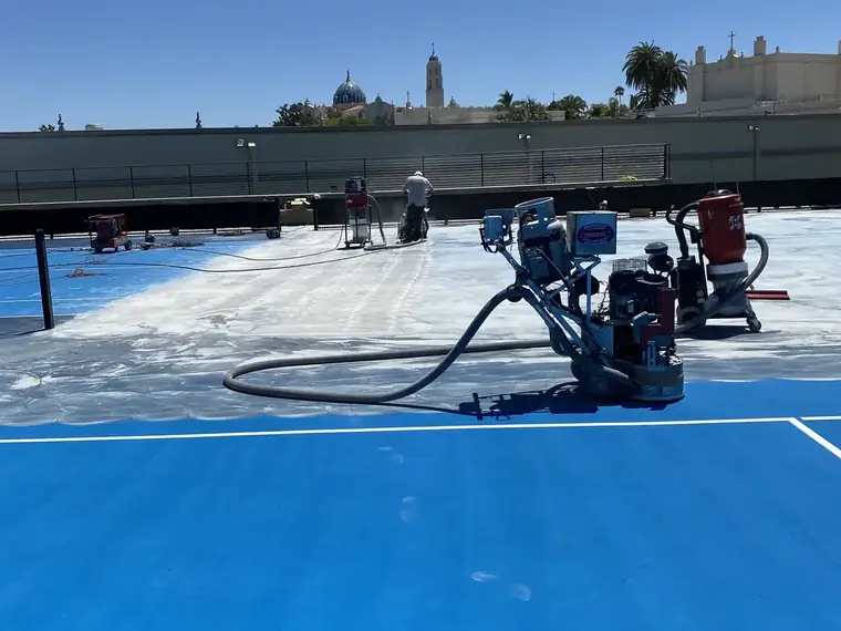 Grinder stripping the blue coating from a court, bare concrete showing behind