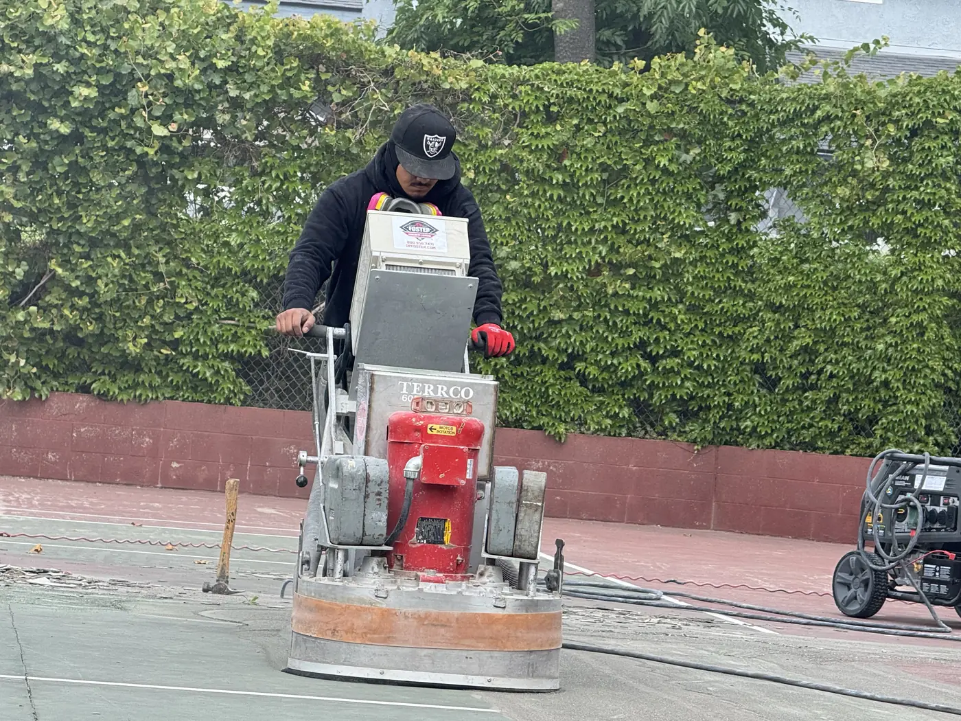 Worker guiding a red walk-behind grinder across a court surface