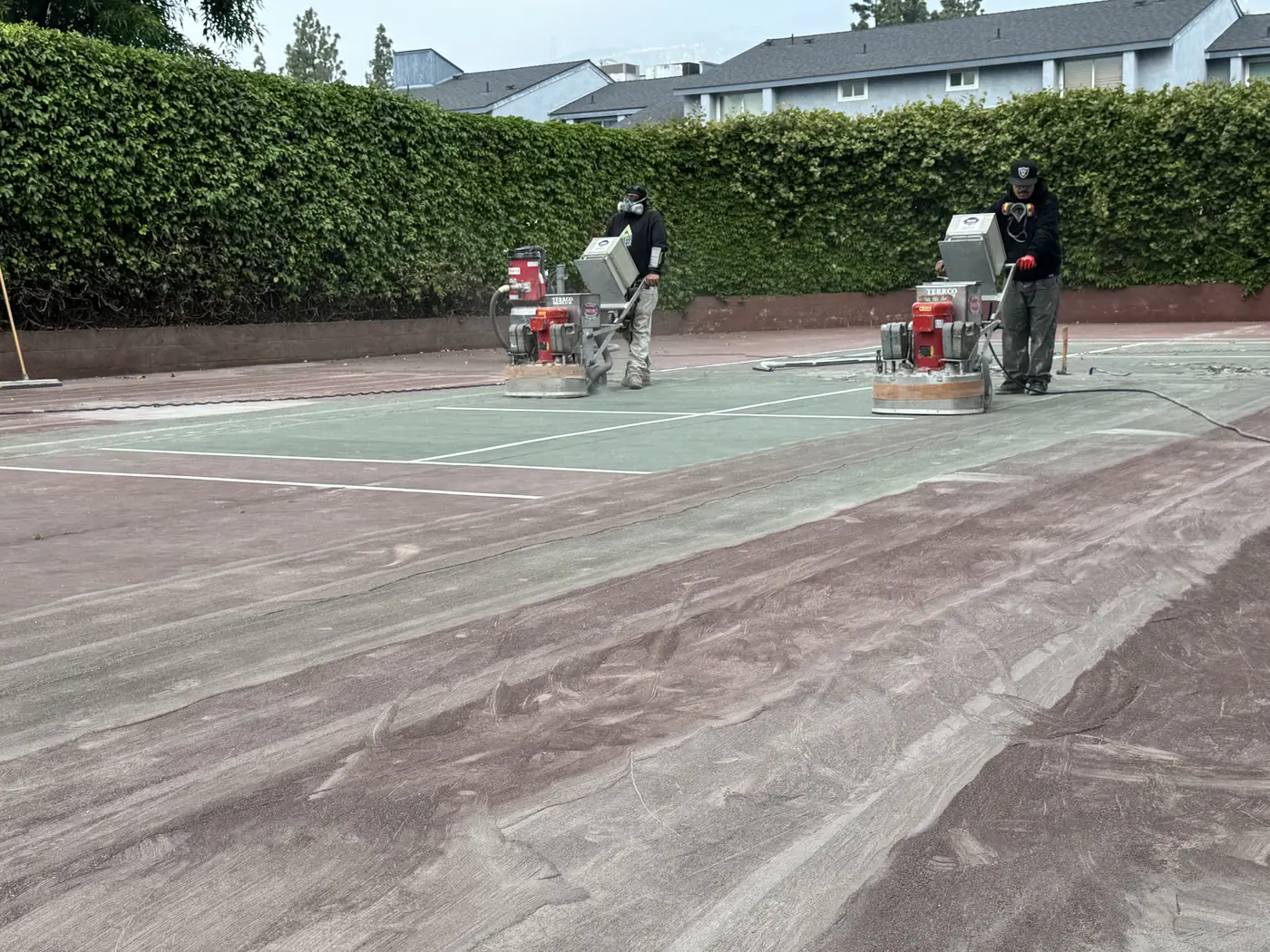 Grinding machines working across a green court beside a tall hedge