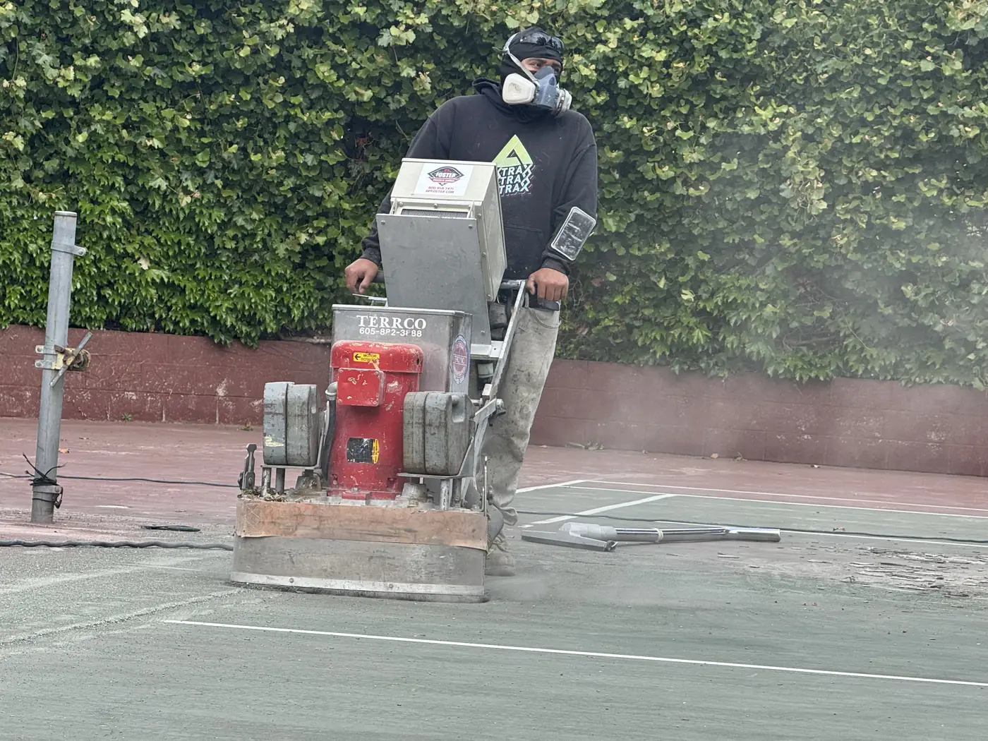 Worker in a respirator running a walk-behind grinder across a green tennis court