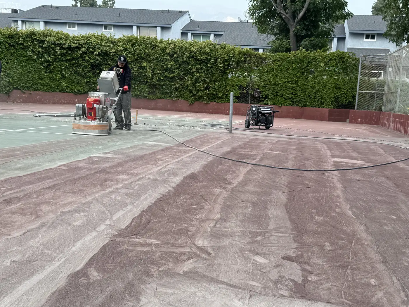 Worker grinding the red surface off a tennis court in front of apartments
