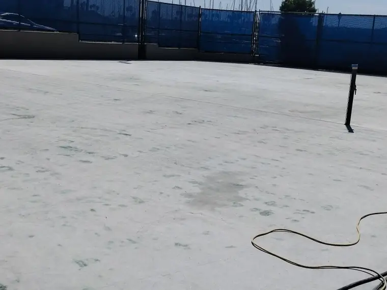 Bare concrete court after the coating was removed, fenced with blue windscreen
