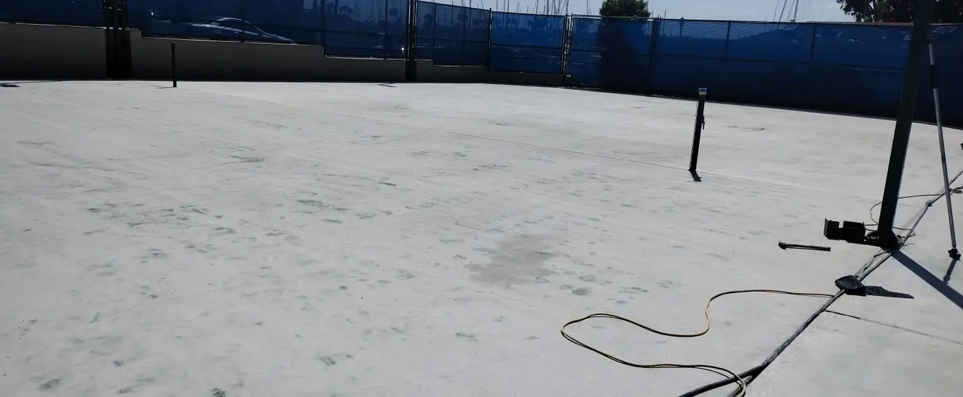 Bare concrete court after the coating was removed, fenced with blue windscreen