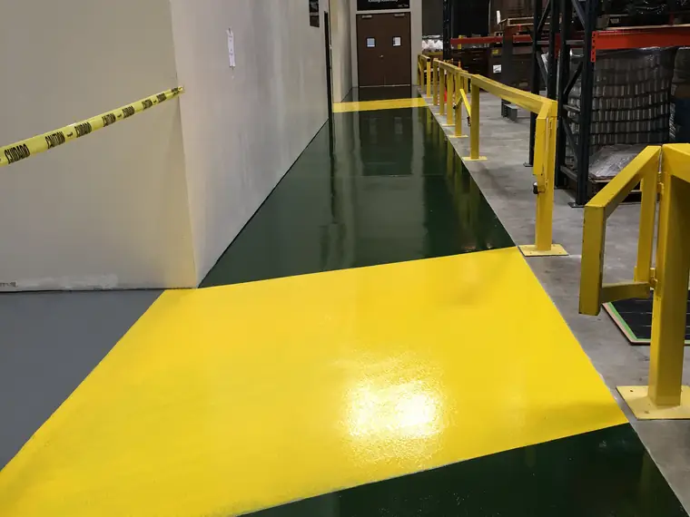 Green and yellow epoxy walkway meeting gray floor coating beside yellow guard rails