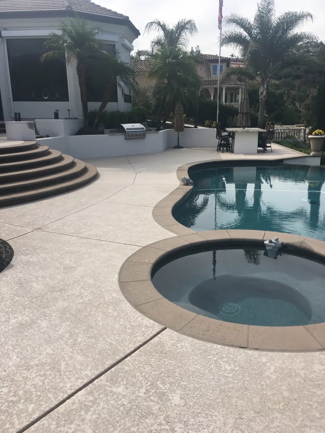 Backyard pool and round spa with a gray textured deck and outdoor kitchen behind