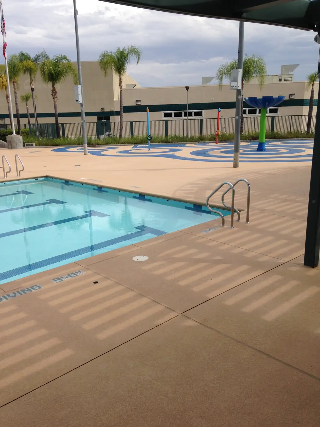 Community pool and splash pad with a tan textured deck and palm trees