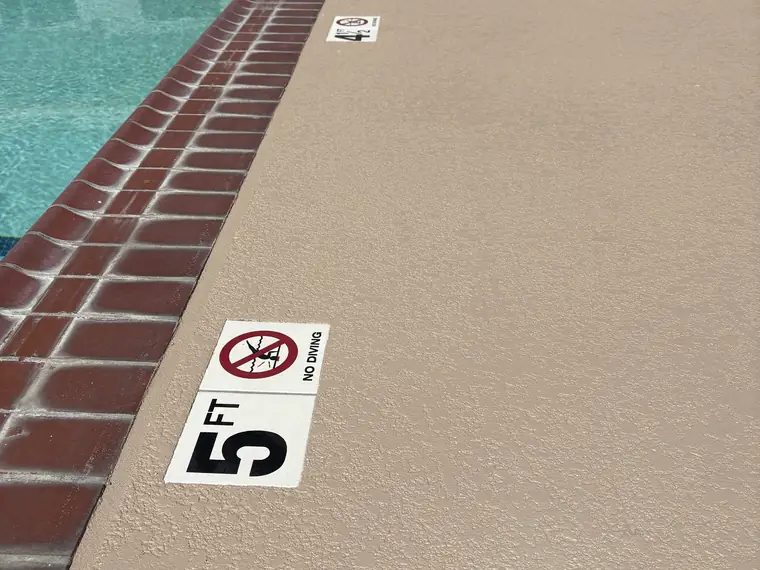 Tan pool deck with a five foot depth marker beside red brick coping