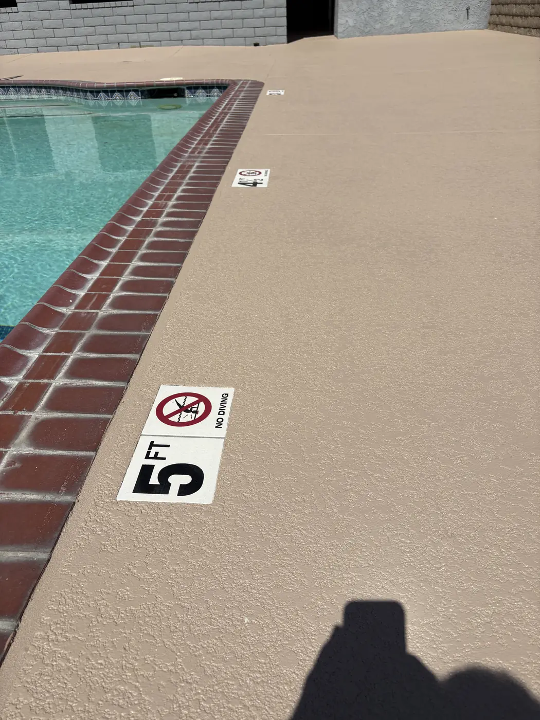Tan pool deck with a five foot depth marker beside red brick coping