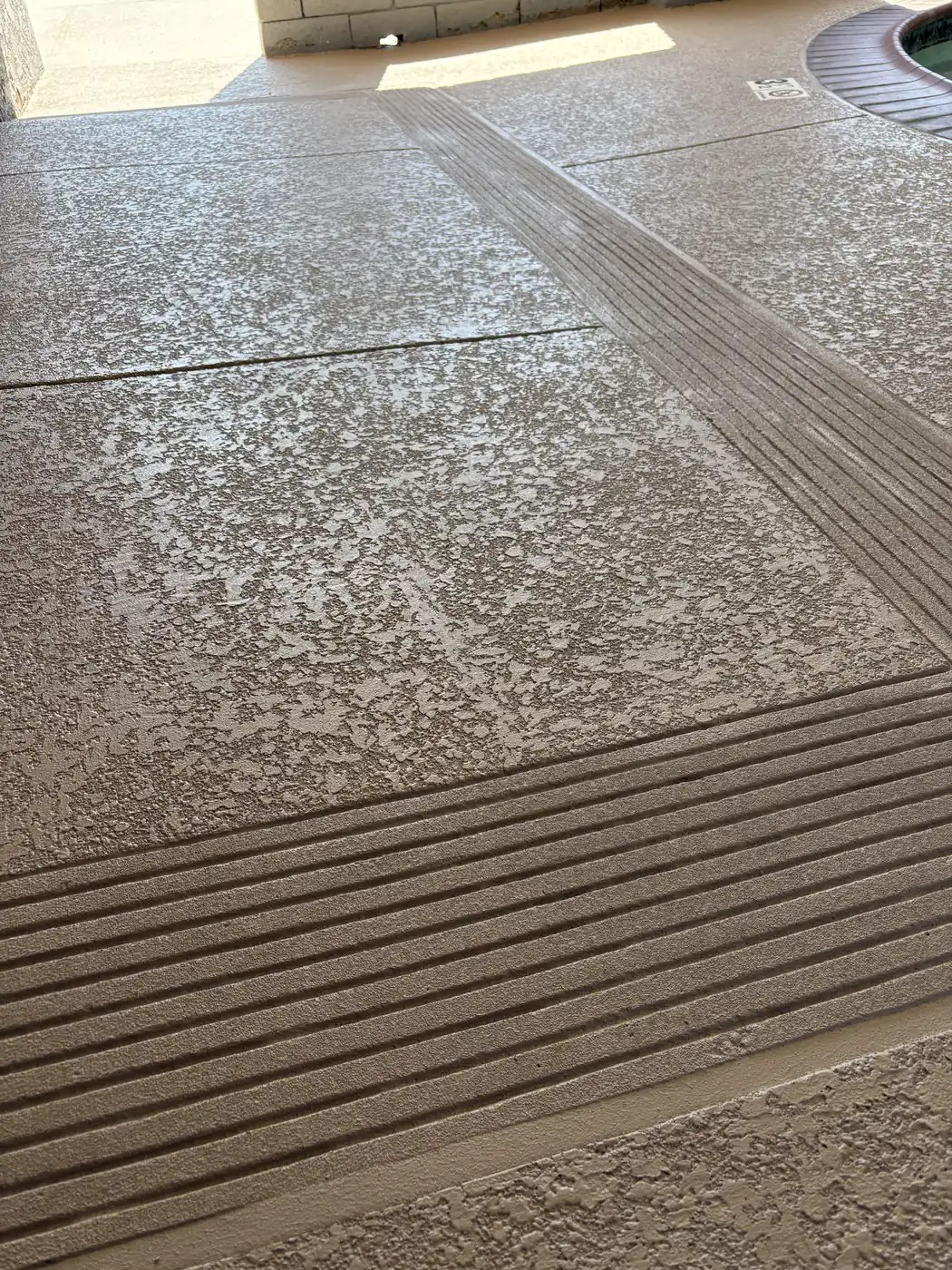 Tan knockdown-textured deck coating with bands of grooved non-slip treads along the step edges, beside a spa under a covered patio