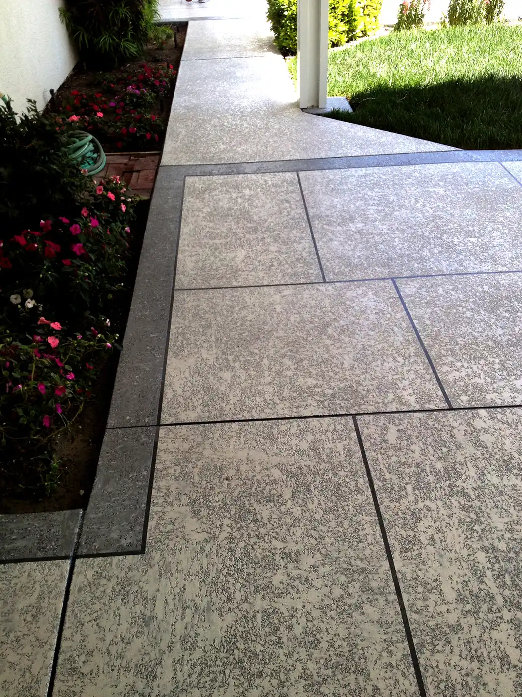 Gray textured walkway with a dark border running past flowering beds