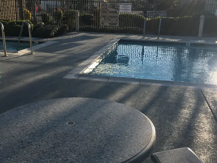 Concrete table and stools on a gray textured pool deck with a blue-gray border beside a community pool