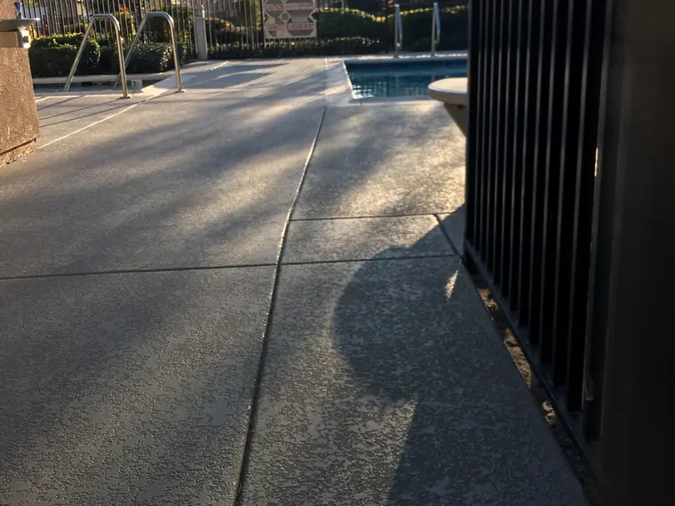 Gray resurfaced walkway with a blue-gray edge band leading past a black fence to a pool