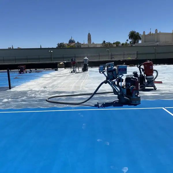 Grinder stripping the blue coating from a court, bare concrete showing behind