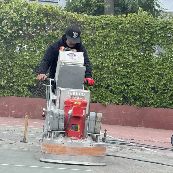 Worker guiding a red walk-behind grinder across a court surface