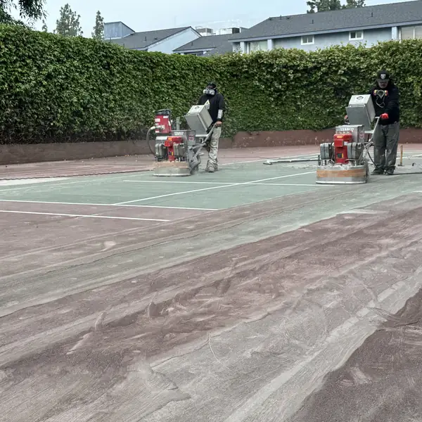 Grinding machines working across a green court beside a tall hedge