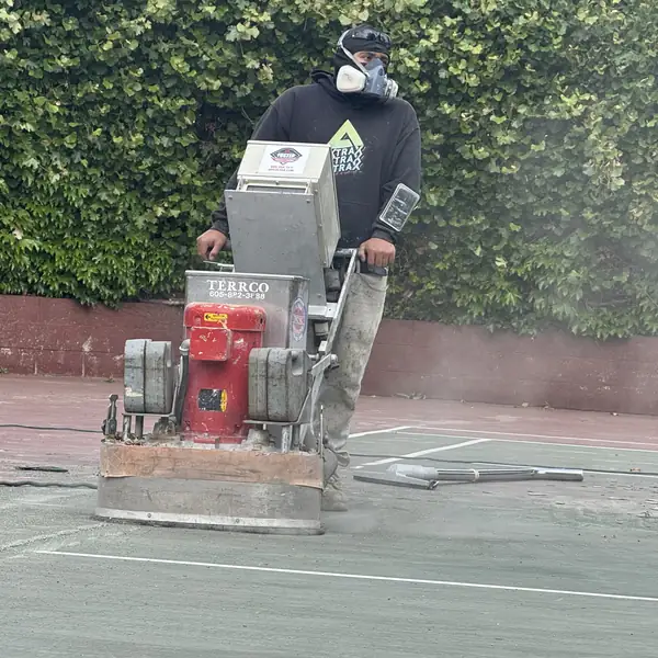 Worker in a respirator running a walk-behind grinder across a green tennis court