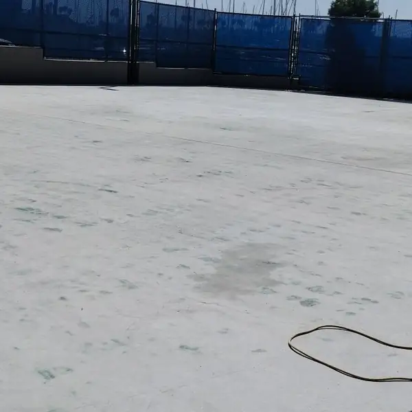 Bare concrete court after the coating was removed, fenced with blue windscreen