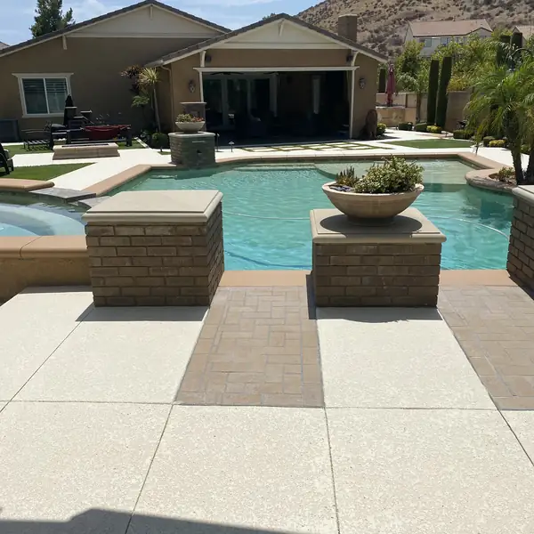 Wide backyard pool with a cream deck and brick planter walls behind a family home