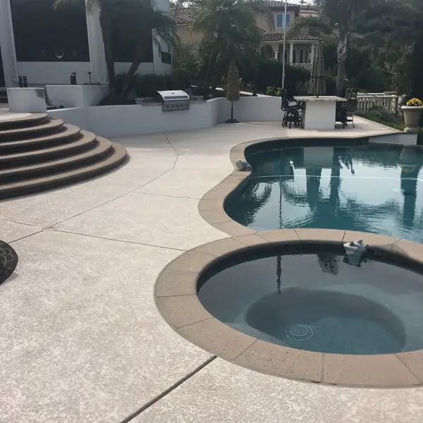 Backyard pool and round spa with a gray textured deck and outdoor kitchen behind
