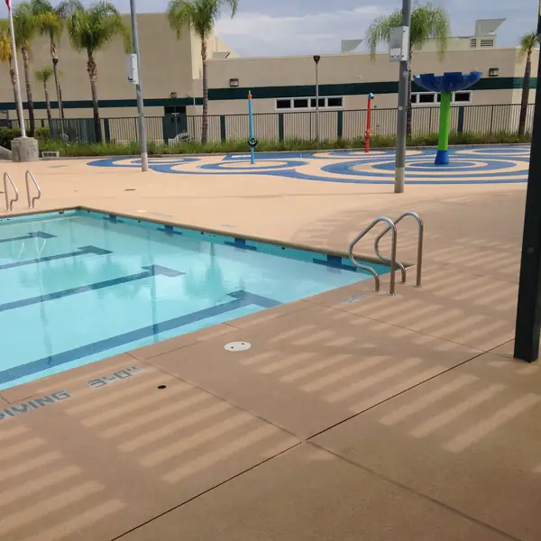 Community pool and splash pad with a tan textured deck and palm trees